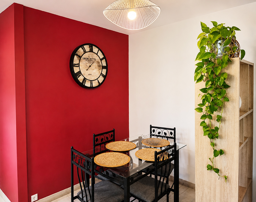 Coin repas avec table en verre, quatre chaises et grande horloge sur un mur rouge.
