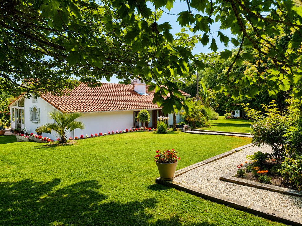 Maison au toit de tuiles bordée d’une pelouse, de fleurs et d’une allée de gravier.